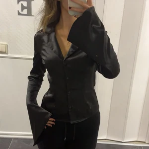 Svart satinblus med knappar - Säljer en svart satinblus med knappar framtill och klassisk krage. Blusen har lång ärm och en elegant, glansig finish som ger en lyxig känsla. Perfekt för dig som vill ha en stilren och trendig look. Passar s också