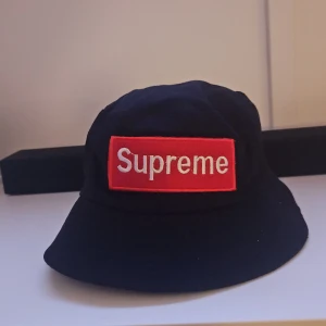 svart bucket hat med röd logga - bucket hat i svart med klassisk röd box-logo framtill. Hatten har bred brätte och är tillverkad i mjuk bomull som ger en chill streetwear-vibe. Perfekt för dig som vill sticka ut med en ikonisk accessoar.