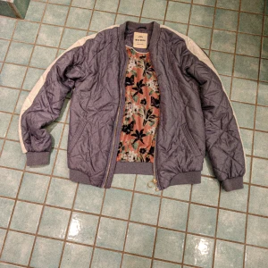 Quiltad bomberjacka av Moss Copenhagen - Helt ny och sjukt snygg ljuslila bomberjacka från Moss Copenhagen med quiltad yta och vita ränder längs ärmarna. Insidan har ett blommigt foder i rosa och svart. Jackan har dragkedja framtill och ribbade muddar vid ärmslut och nederkant.