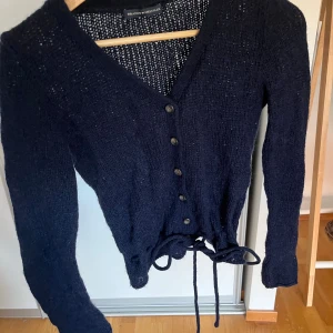 Mörkblå stickad kofta Brandy Melville - Mysig mörkblå stickad kofta från Brandy Melville med v-ringning, knappar framtill och snörning i midjan. Långärmad och perfekt att slänga på sig när det är lite kyligare ute.