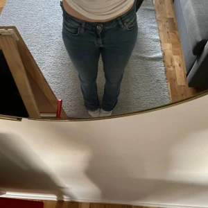 Blå straight jeans från Zara, strl 34 - Säljer ett par klassiska blå jeans från Zara i storlek 34. Modellen har normal midja och bootcut. Jeansen är tillverkade i jeansmaterial och har fem fickor samt knapp och dragkedja framtill. Perfekt passform för dig som gillar enkel och tidlös stil.