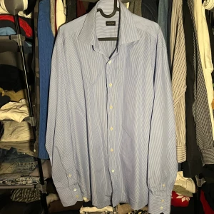 Blårandig skjorta från Valentino Roma - Snygg skjorta från Valentino Roma i ljusblått med tunna vita ränder. Klassisk krage, långärmad och med knappar framtill samt vid manschetterna. Skjortan har en loose passform och är tillverkad i mjuk bomull som känns skön mot huden. Perfekt för dig som gillar stilrena och tidlösa plagg.