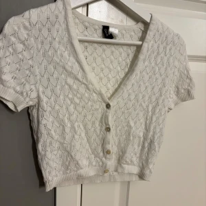 Vit stickad blus från DIVIDED - Supersöt vit stickad blus från DIVIDED i storlek S. Den har korta ärmar, v-ringning och knappar framtill. Mönstret är diskret och ger en fräsch känsla, perfekt för vår och sommar. Ribbad kant vid ärmslut och nederkant för extra stilpoäng.