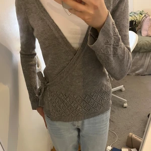 Grå omlottkofta med knytband och fint mönster - Koftan har ett snyggt hålmönster med diamantformade detaljer både fram och bak samt nedtill. Lite smått utsvängda ärmar. Skriv för fler bilder eller info!