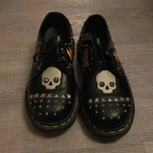 Dr. Martens svarta loafers med nitar och döskalle - Unika svarta Dr. Martens loafers med coola döskallebroderier, nitar och patchar som blixtar och brittiska flaggan. Skorna har gul söm runt sulan och är tillverkade i skinn. Perfekta för dig som vill sticka ut med en edgy stil.