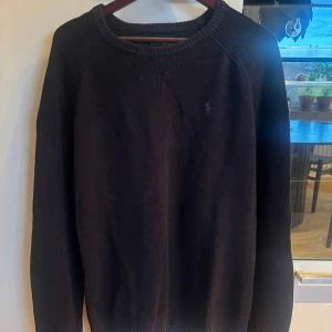 Svart stickad tröja från Polo Ralph Lauren - Klassisk svart stickad tröja från Polo Ralph Lauren med diskret broderad logga på bröstet. Tröjan har rund halsringning, ribbade muddar och lång ärm. Perfekt för lager-på-lager och enkel att matcha med jeans eller chinos.