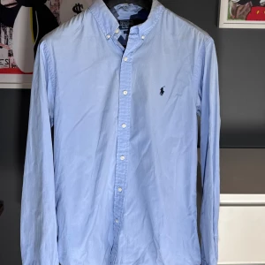 Ljusblå skjorta Polo Ralph Lauren M - Nu är denna eftertraktade och snygga Ralph Lauren skjorta till salu. En väldigt stilren och simpel skjorta i size M (slim fit).  Cond 8,5/10 (skjortan är lite mindre i storleken)  Nypris: 1599 kr - Vårt pris 599 kr.  Endast skjorta som medföljer vid köp.  Snabba och enkla affärer prioriteras i vanlig ordning. 