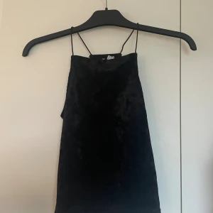 Svart sammetstopp med tunna band - Supersnygg svart kort sammetstopp med tunna axelband och rak halsringning. Toppem har en enkel, stilren design och är ärmlös vilket gör den perfekt för att styla med smycken eller en cool jacka. Passar dig som gillar en klassisk men trendig look.
