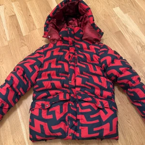 Vinterjacka från J.Lindeberg - Säljer en fet pufferjacka från J.Lindeberg med röd och marinblå grafisk design. Jackan har huva, stora fickor med knappar och dragkedja framtill. Perfekt för kalla dagar när du vill sticka ut med en cool look. Passar S & M