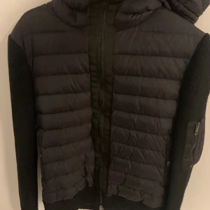 Svart Moncler Cardigan helt nyskick 10/10 - Svart pufferjacka från Moncler med stickade ärmar och huva. Jackan har quiltad framsida, dragkedja och en ficka med dragkedja på ärmen. Klassisk Moncler-logga på vänster ärm. Perfekt för kyliga dagar med sin kombination av dun och stickat material.