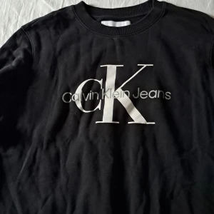 Svart sweatshirt från Calvin Klein Jeans - Svart sweatshirt från Calvin Klein Jeans med stor vit CK-logga och text på bröstet. Klassisk rund halsringning och ribbade muddar. Tillverkad i mjuk bomullsmix som är skön att bära. Perfekt för en stilren och avslappnad look.