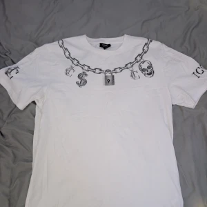 Vit t-shirt ICONO - Vit t-shirt från ICONO med tryck av kedja, hänglås, dollartecken och dödskallar runt halsen och på ärmarna. Regular fit med klassisk rund hals och korta ärmar. Skön och lätt i bomull, perfekt för en edgy streetstil. Strl L skriv för frågor, bilder och offer (: