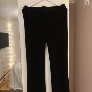 Svarta kostymbyxor från Vero Moda - Snygga svarta kostymbyxor från Vero Moda med klassisk rak passform. Byxorna har hög midja, bälteshällor, dragkedja och både knapp och hake i linningen. Perfekta för en clean och stilren look.