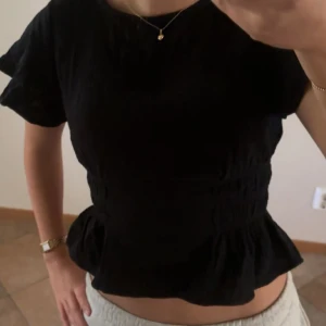 Svart blus med volang - Svart blus med korta ärmar och volang nertill. Modellen har en lätt rynkad midja som ger en feminin siluett. Perfekt att matcha med både jeans och kjol.