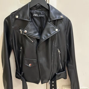  Ny Svart skinnjacka från Zara med bälte - Snygg svart imitation skinnjacka från Zara med klassisk bikerstil, silverfärgade dragkedjor, knappar och bälte i midjan. Jackan har flera fickor med dragkedja och coola detaljer som axelklaffar och bred krage. Perfekt för en edgy look.