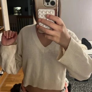 Vit croppad stickad tröja med v-ringning - Säljer en vit croppad stickad tröja med v-ringad hals och långa ärmar. Tröjan har breda ribbade muddar vid ärmslut och nederkant, vilket ger en chill och modern vibe. Perfekt för att styla med jeans eller kjol.