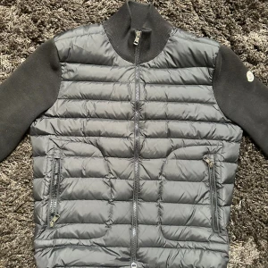 Svart Cardigan från Moncler - Säljer en svart pufferjacka från Moncler med stickade ärmar och hög krage. Jackan har två dragkedjeförsedda fickor framtill och Moncler-logga på ena ärmen. Perfekt för dig som vill ha en stilren och varm jacka med snygga detaljer.