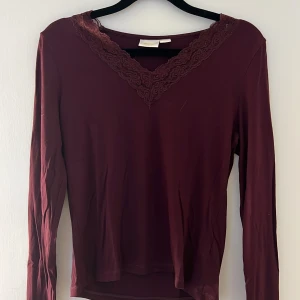 Vinröd långärmad topp från Gina Tricot - Säljer en vinröd långärmad topp från Gina Tricot med v-ringning och spetsdetaljer längs halsen. Toppen är i mjukt material och har en elegant, feminin look som passar perfekt till jeans eller kjol.