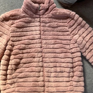 Rosa pälsjacka från b.young - Supermjuk och fluffig rosa pälsjacka från b.young i storlek 36. Jackan har horisontella ränder, hög krage och dragkedja framtill. Perfekt för dig som vill ha en mysig och trendig look med lite extra attityd.