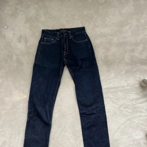Mörkblå jeans från Nudie Jeans - Säljer ett par mörkblå jeans från Nudie Jeans med klassisk femficksdesign och kontrastsömmar. Jeansen har rak passform och är tillverkade i kraftigt denimtyg av bomull. Perfekta för dig som gillar stilrena och tidlösa jeans MODELL: Gritty Jackson. Jeansen är krympta och jag skulle nu säga att dem är i size W28 L30-31. Ursprungligen W29 L32