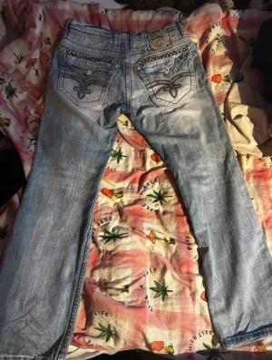 Rock Revival Jeans - Säljer ett par ljusblå jeans från Rock Revival med snygga slitningar och broderade detaljer på bakfickorna. Jeansen har raka ben, klassisk femficksdesign och coola kontrastsömmar längs sidorna. Perfekt för dig som gillar street och unik stil.