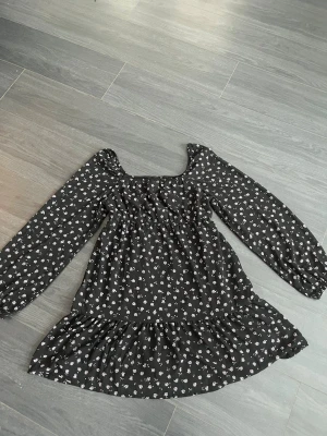 Svart blommig klänning från H&M - Supersöt svart kortklänning från H&M med små lila blommor. Klänningen har långärmade puffärmar, markerad under bysten  och volang nertill. Tillverkad i mjuk polyester, perfekt för dig som gillar romantiska och trendiga vibes.
