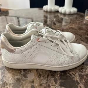 Vita sneakers från Björn Borg - Snygga vita sneakers från Björn Borg med perforerade detaljer på sidorna och metallicfärgad häl. Klassisk design med snörning och diskret logga på sidan. Perfekta för en clean och sportig look.
