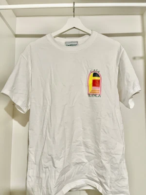  Casablanca t-shirt  - Casablanca t-shirt i vitt med korta ärmar och rund hals. Framtill och baktill finns en stor grafisk print i rosa, gul, röd, orange och svart med texten CASA BLANCA. Materialet är mjuk bomull och passformen är normal. Perfekt för dig som gillar snygga prints och streetwear.