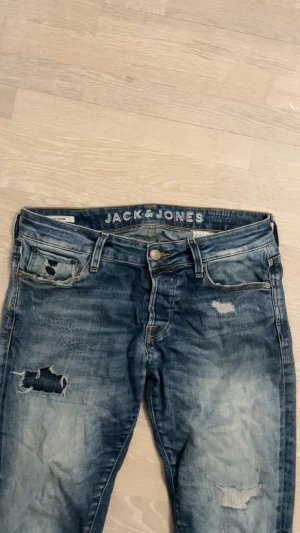 Blå slitna jeans från Jack & Jones - Säljer ett par blå jeans från Jack & Jones med snygga slitningar och hål på benen för en cool, avslappnad look. Jeansen har klassisk femficksdesign, knappgylf och brunt läderpatch bak i midjan. Passformen är slim. Jeansen har modellen Glenn. Storleken är W32 och L30