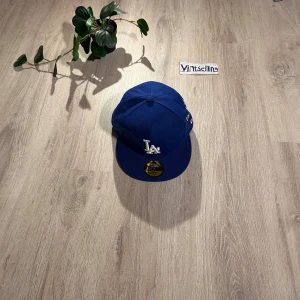 Blå LA Dodgers keps från New Era - Snygg blå LA Dodgers keps från New Era 59FIFTY med vit broderad LA-logga framtill och cool textbrodyr på sidan och bak. Klassisk flat peak och MLB-märke baktill. Perfekt för dig som gillar streetstyle och sportig vibe.
