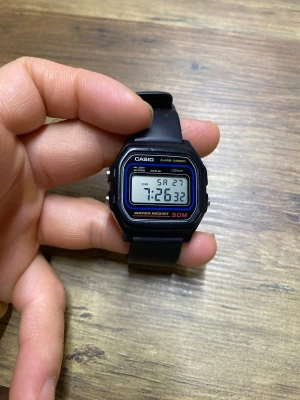 Svart digital Casio klocka 50M - Klassisk digital Casio klocka med svart fyrkantigt plastboett och svart gummiband. LCD-display med tid, datum och alarmfunktion. Vattentät upp till 50 meter och har silverfärgade knappar på sidan. Perfekt för dig som gillar retro och funktion.