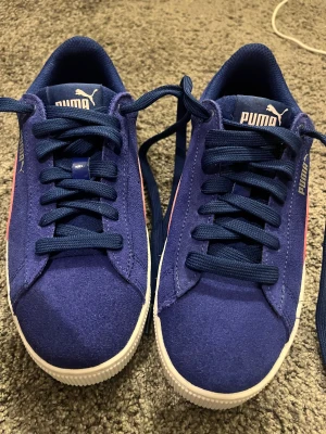 Blå och rosa sneakers från Puma - Snygga sneakers från Puma i marinblå mocka med klassisk rosa sidostrip och vit sula. Skorna har rund tå, platt sula och snörning. Perfekta för dig som vill ha en sportig och trendig look med en färgklick.