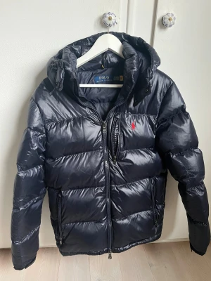 Mörkblå pufferjacka Polo Ralph Lauren - Snygg mörkblå pufferjacka från Polo Ralph Lauren med huva och dragkedja. Jackan har klassisk quiltad design, två sidofickor med dragkedja och Polo-logga i rött på bröstet. Har använts sparsamt och har endast en liten defekt som man kan se på bild 4. Går fortfarande att hänga den. Kontakta vid minsta fundering!
