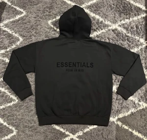 Svart Essentials Fear of God hoodie - Svart hoodie från Essentials Fear of God med stor logga på ryggen och diskret patch på framsidan. Klassisk huva och magficka, tillverkad i mjuk bomull. Perfekt för dig som gillar streetwear och vill ha en clean look.
