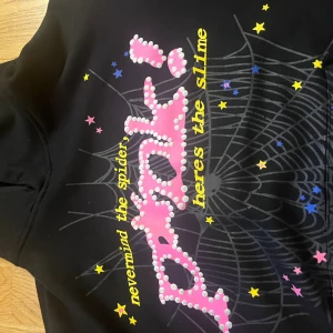 Sp5der p*nk!  - Svart hoodie med stor huva och coolt tryck på framsidan med texten 'nevermind the spider, heres the slime', rosa och vita prickar, stjärnor i gult, blått och rosa samt spindelnätsmönster. På ryggen finns en gul stjärna med ST★R-logga. Perfekt för dig som gillar unika streetwear-plaggp. Den är lite söder längst framme det är därför den e billig 