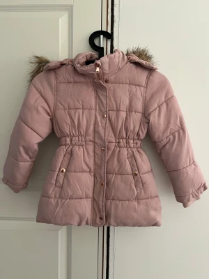 Rosa pufferjacka med fuskpäls KappAhl 116 - Supersöt rosa pufferjacka från KappAhl med mysig fuskpäls på huvan och fleecefoder. Jackan har dragkedja, knappar, två fickor och en söt reflexdetalj med katt på ryggen. Perfekt för kalla dagar och riktigt skön att ha på sig.