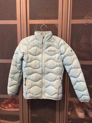 Ljusblå dunjacka från Peak Performance - Säljer en ljusblå dunjacka från Peak Performance med vågquiltat mönster och hög krage. Jackan har dragkedja framtill, två fickor med dragkedja och logga på bröstet. Perfekt för kyliga dagar och riktigt snygg till streetwear.