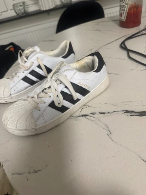 Adidas Superstar vita sneakers - Klassiska Adidas Superstar sneakers i vitt med svarta ränder och guldtext på sidan. Skorna har snörning, platt sula och den ikoniska rundade tån med mönstrad gummidetalj. Tillverkade i skinn och syntetmaterial, passar perfekt för dig som gillar retrostil.