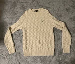 Beige stickad tröja från Lyle & Scott - Snygg beige stickad tröja från Lyle & Scott med klassiskt kabelmönster och gul logotyp på bröstet. Rund hals och ribbade muddar vid ärmslut och nederkant. Perfekt för dig som gillar stilrena och tidlösa plagg.