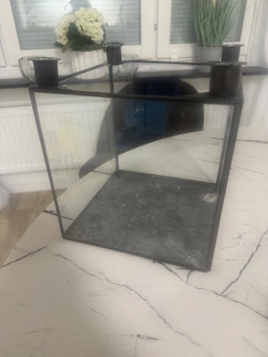 Ljuhålare  - Bilden visar en fyrkantig glaslykta med metallram och plats för fyra värmeljus på toppen. Produkten har viss patina och spår av användning på bottenplattan. Perfekt som dekorativ ljushållare för hemmet. Ingen elektronik synlig.