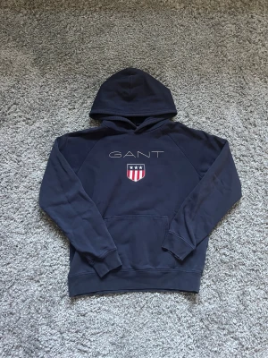 Marinblå hoodie från GANT - Snygg marinblå hoodie från GANT med broderad logga och emblem på bröstet. Tröjan har huva, känguruficka och långa ärmar. Tillverkad i mjuk bomullsmix som är skön att ha på sig. Perfekt för dig som gillar klassisk och sportig stil.