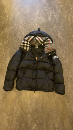 Svart dunjacka Burberry med avtagbara ärmar - Svart pufferjacka från Burberry med avtagbara ärmar så du kan bära den som väst. Jackan har klassiskt rutigt foder i huvan, dragkedja och knappar framtill samt två fickor med dragkedja. Märkespatch på ena ärmen och snygg, boxig passform.