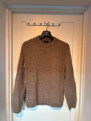 Beige stickad tröja från GANT - Säljer en beige stickad tröja från GANT i storlek S. Tröjan har grov ribbstickning, rund hals och diskret broderad logga på bröstet. Perfekt för dig som gillar klassisk och chill stil med lite extra textur.