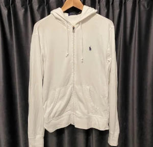 Vit hoodie från Polo Ralph Lauren - Vit hoodie från Polo Ralph Lauren med klassisk broderad logga på bröstet. Tröjan har huva med snörning, två fickor fram och dragkedja med Polo-detalj. Långärmad och tillverkad i mjuk bomull, perfekt för chill dagar.
