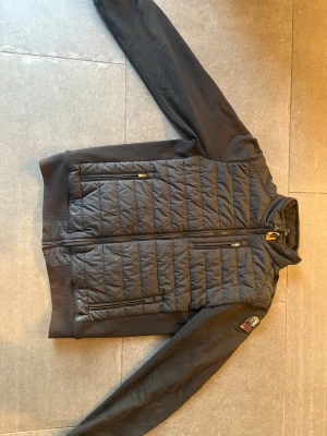 Parajumpet cardigan storlek Y-L - Snygg svart pufferjacka från Parajumpers med quiltad framsida och rygg, samt släta ärmar. Jackan har dragkedja framtill, två dragkedjefickor och en patch på ärmen. Insidan är fodrad i silver och har en säkerhetsficka. Perfekt för kyliga dagar. Ungdoms storlek 