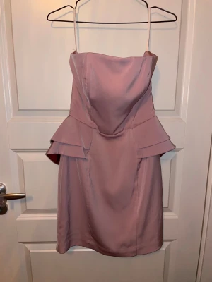 Axelbandslös rosa kort klänning med volang - Snygg axelbandslös kort klänning i ljusrosa med volangdetaljer vid midjan. Klänningen har en figurnära passform och dragkedja bak. Materialet är slätt och har en lätt glans, perfekt för dig som vill sticka ut med en feminin look.