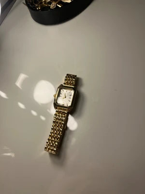 Guldfärgad fyrkantig klocka  - Snygg guldfärgad klocka från Axiver med fyrkantigt ur och länkarmband i metall. Urtavlan är silverfärgad med minimalistiska guldmarkeringar och visare. Länken har en klassisk design som ger en lyxig känsla och passar perfekt till dig som gillar stilrena accessoarer.