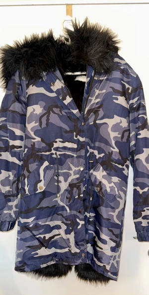 Super snygg/ varm Blå/grå camo parkas från NLY Trend ( Nelly ) - Oversized- Parkasjacka i blått och grått camouflagemönster från NLY Trend. Jackan har stor huva och foder i svart fuskpäls, dragsko i midjan och två stora fickor framtill. Perfekt för kalla dagar när du vill sticka ut lite extra.