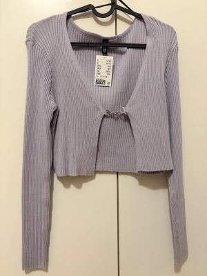 Ljuslila ribbad croppad bolerotopp H&M - Superfin ljuslila croppad bolerotopp från H&M med ribbad struktur. Toppen har lång ärm, v-ringning och en snygg silverfärgad hake framtill som stängning. Perfekt att styla över linne eller klänning för en trendig look.