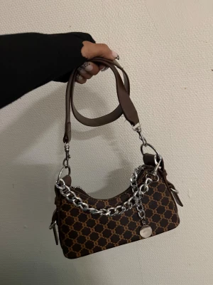 Brun handväska med kedjedetalj - Snygg brun handväska med grafiskt mönster och silverfärgad kedja framtill. Väskan har ett avtagbart handtag i brunt skinnliknande material och dragkedja upptill. Perfekt storlek för det viktigaste och en cool detalj till din outfit.